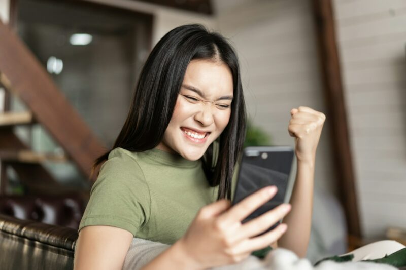 Winning asian girl sitting at home and looking at mobile phone, cheering and celebrating from good