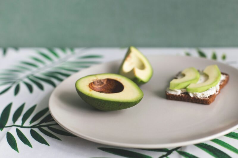 On a plate is a ripe avocado and a sandwich of cereal bread, soft cheese and slices of avocado