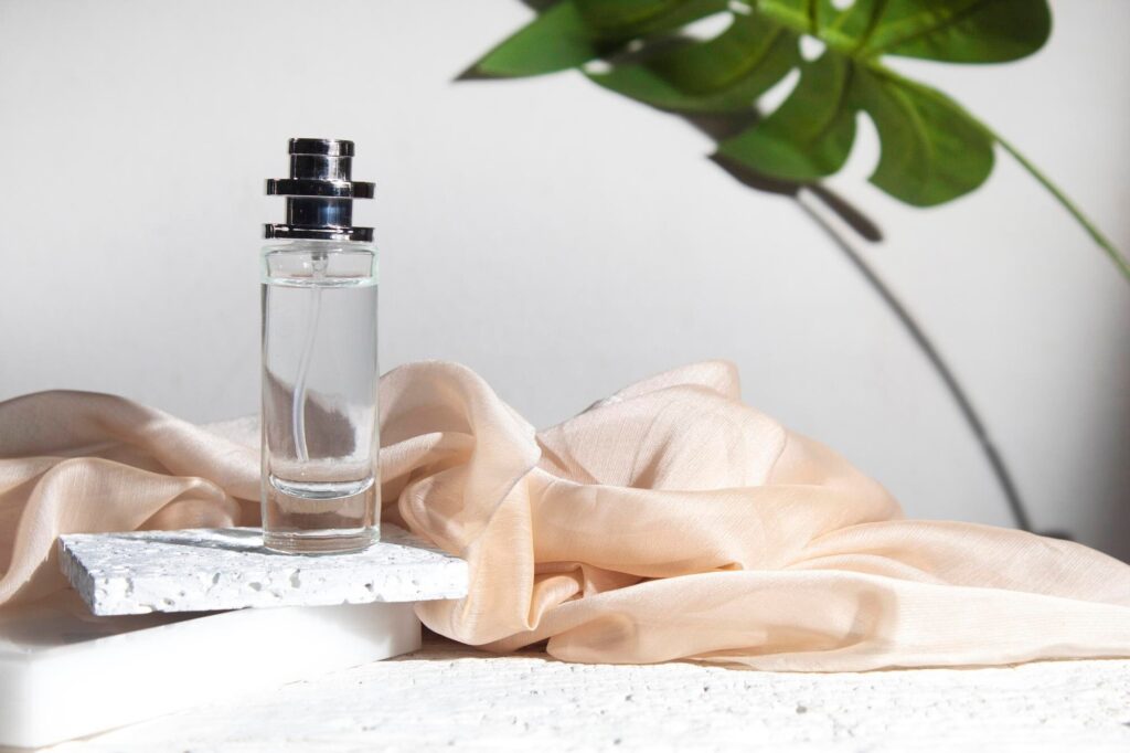 Frasco de perfume de vidro transparente com tampa prateada sobre placa de pedra, ao lado de tecido bege e folha verde