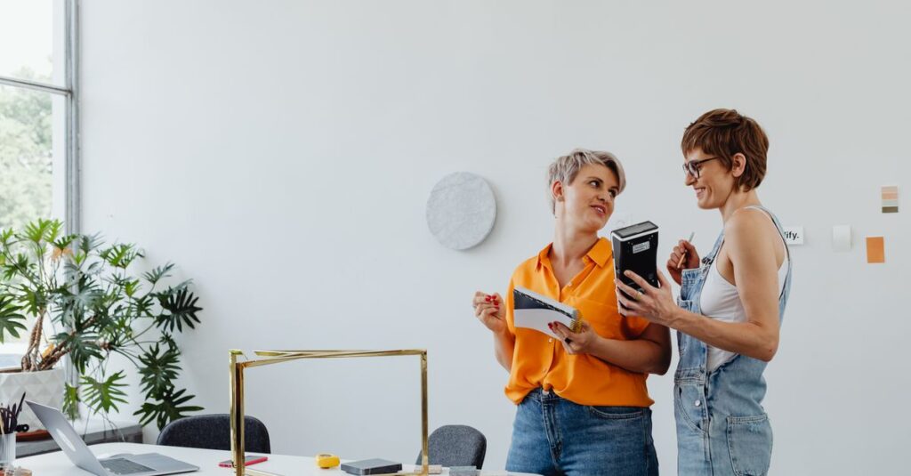 Como transformar estruturas metálicas em escritórios modernos e funcionais Duas mulheres, uma com blusa laranja e outra com macacão, conversando em um espaço de trabalho iluminado.