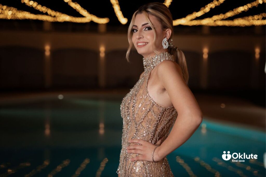 Como montar looks de moda para vida noturna feminina Mulher sorridente usando vestido de festa com detalhes brilhantes próximo a uma piscina iluminada.