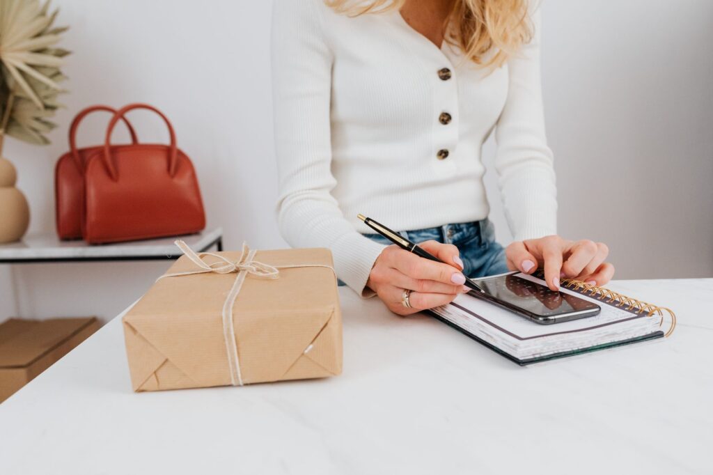 Frete caro faz 57% dos brasileiros abandonarem compras online Mulher confere o celular ao lado de uma encomenda embrulhada em papel kraft sobre a mesa
