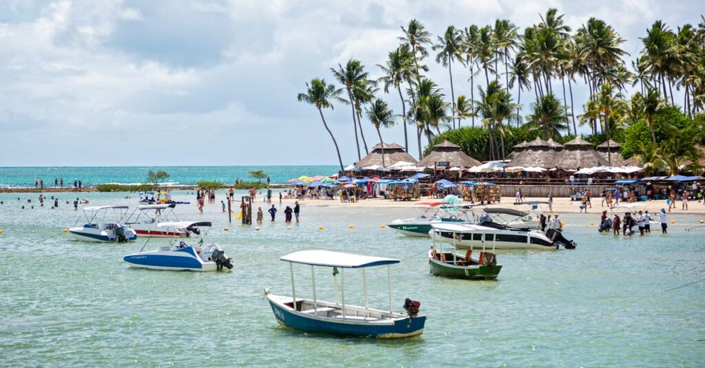 Vista de uma praia com pessoas, barcos e palmeiras sob um céu azul.