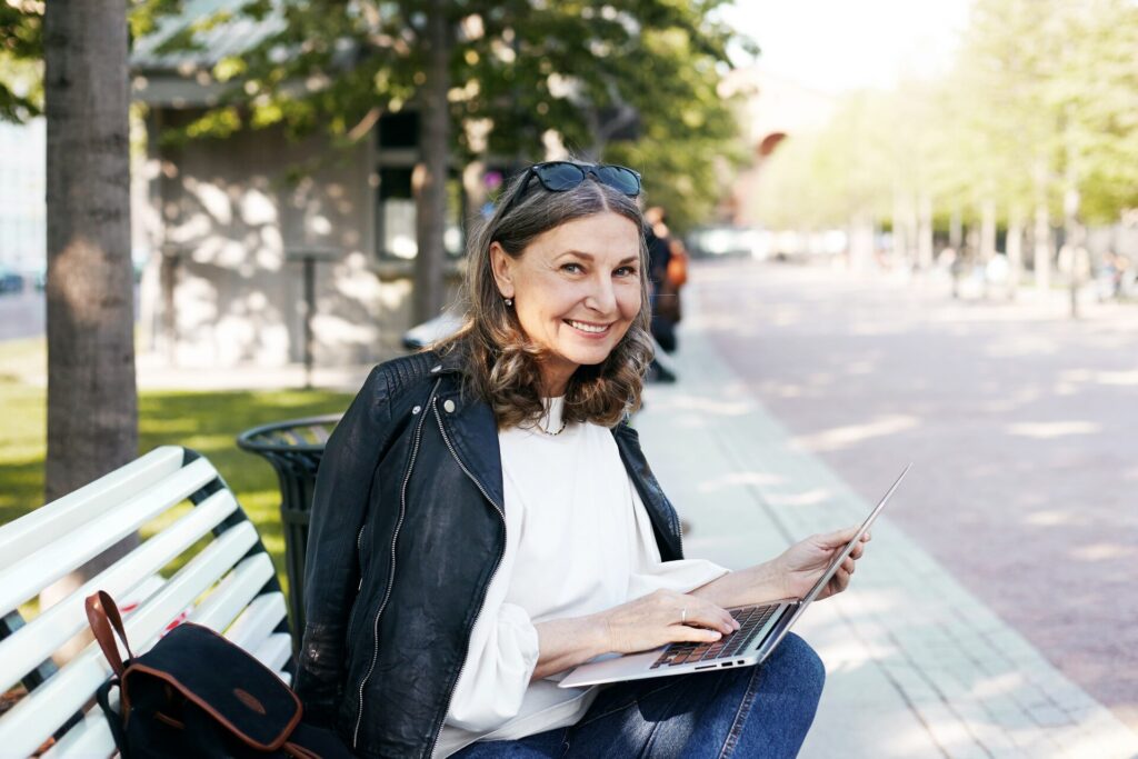 Mulher sênior sorridente sentada em banco, usando laptop